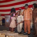 A U.S. Navy promotion or advancement ceremony is taking place in front of a large American flag. A sailor in khaki dress uniform is kneeling while a young girl pins insignia or a device onto his collar, surrounded by family members and fellow naval personnel. Engraved wooden plaques or cases are visible in the foreground, indicating this is a formal recognition event.