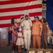 A U.S. Navy promotion ceremony is taking place in front of a large American flag backdrop. A sailor in khaki uniform is having his chief petty officer cover (hat) placed on his head simultaneously by family members, a traditional 'pinning' or 'capping' ritual. Family members including a woman, a young girl, and an older couple participate alongside a senior naval officer observing from the right.