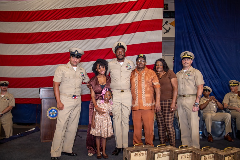 A U.S. Navy promotion or chief petty officer pinning ceremony is taking place in front of a large American flag. A newly promoted sailor in khaki chief's uniform poses with family members and two flanking naval officers. Engraved wooden plaques or boxes are visible in the foreground, consistent with a formal military advancement ceremony.