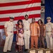 A U.S. Navy promotion or chief petty officer pinning ceremony is taking place in front of a large American flag. A newly promoted sailor in khaki chief's uniform poses with family members and two flanking naval officers. Engraved wooden plaques or boxes are visible in the foreground, consistent with a formal military advancement ceremony.