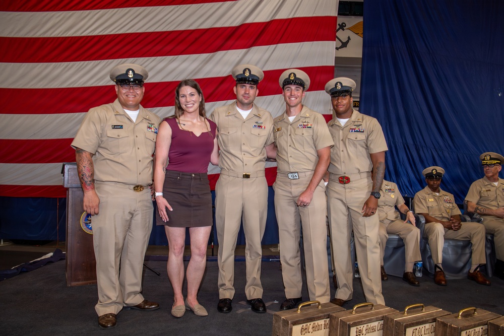 A group photo of four U.S. Navy personnel in khaki service uniforms and one civilian woman posing together in front of a large American flag backdrop. The setting appears to be a formal military ceremony or promotion event, with additional uniformed personnel seated in the background. Several name placards or award cases are visible in the foreground.