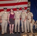 A group photo of four U.S. Navy personnel in khaki service uniforms and one civilian woman posing together in front of a large American flag backdrop. The setting appears to be a formal military ceremony or promotion event, with additional uniformed personnel seated in the background. Several name placards or award cases are visible in the foreground.