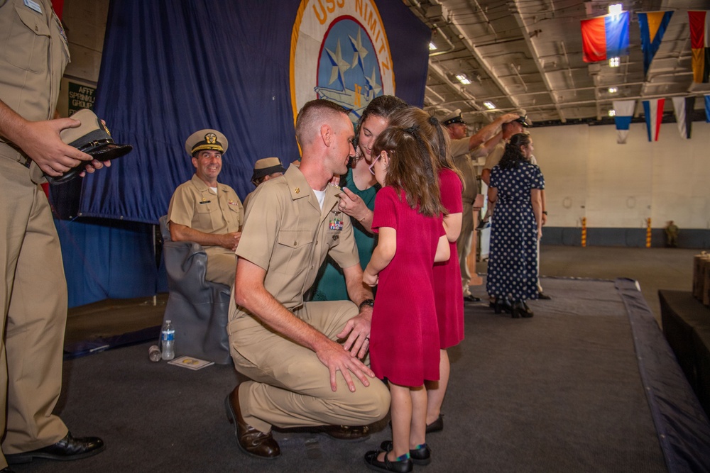 A U.S. Navy sailor in service khaki uniform kneels down to receive a kiss or pin from a young girl in a red dress, while a woman in green stands nearby during what appears to be a military promotion or award ceremony. The event is taking place aboard the USS Nimitz, as indicated by the large blue banner in the background. Other naval personnel and civilians are visible in the background observing the ceremony.