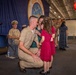 A U.S. Navy sailor in service khaki uniform kneels down to receive a kiss or pin from a young girl in a red dress, while a woman in green stands nearby during what appears to be a military promotion or award ceremony. The event is taking place aboard the USS Nimitz, as indicated by the large blue banner in the background. Other naval personnel and civilians are visible in the background observing the ceremony.