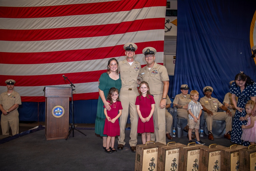 A U.S. Navy military ceremony, likely a promotion or chief petty officer pinning ceremony, taking place in a decorated hall with a large American flag as backdrop. Two Navy personnel in khaki uniforms pose with a woman in a green dress and two young girls in matching red dresses, with engraved wooden plaques visible in the foreground. Additional Navy personnel, families, and children are visible in the background.