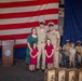 A U.S. Navy military ceremony, likely a promotion or chief petty officer pinning ceremony, taking place in a decorated hall with a large American flag as backdrop. Two Navy personnel in khaki uniforms pose with a woman in a green dress and two young girls in matching red dresses, with engraved wooden plaques visible in the foreground. Additional Navy personnel, families, and children are visible in the background.