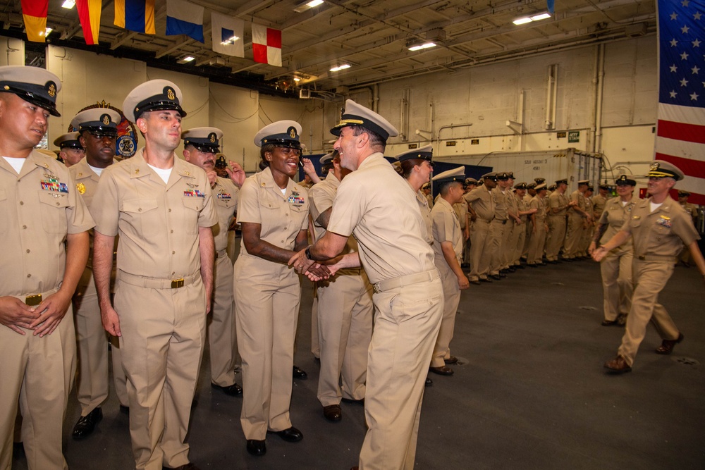 A U.S. Navy ceremony is taking place inside what appears to be a ship's hangar bay, with personnel in khaki service uniforms lined up in formation. A senior officer is moving down the line shaking hands with sailors, suggesting a recognition, promotion, or change of command ceremony. An American flag and various naval signal flags are visible in the background.