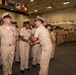 A U.S. Navy ceremony is taking place inside what appears to be a ship's hangar bay, with personnel in khaki service uniforms lined up in formation. A senior officer is moving down the line shaking hands with sailors, suggesting a recognition, promotion, or change of command ceremony. An American flag and various naval signal flags are visible in the background.