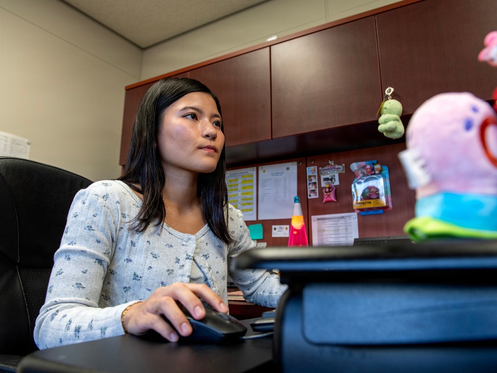 A young woman with dark hair is seated at an office desk, using a computer mouse while looking at a monitor off-frame. The workspace features a bulletin board with pinned documents, a lava lamp, and colorful plush toys on the desk. The setting appears to be a small, personalized office cubicle or private office.