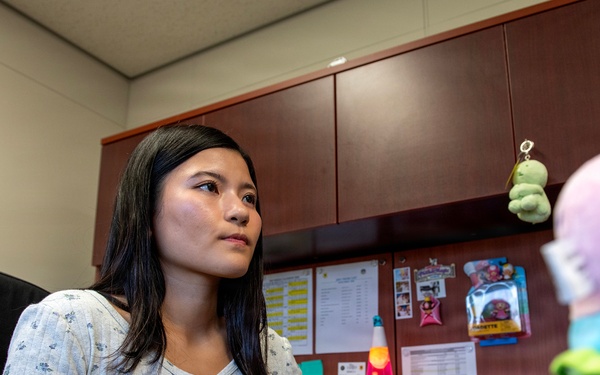 A young woman with dark hair is seated at an office desk, using a computer mouse while looking at a monitor off-frame. The workspace features a bulletin board with pinned documents, a lava lamp, and colorful plush toys on the desk. The setting appears to be a small, personalized office cubicle or private office.