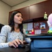 A young woman with dark hair is seated at an office desk, using a computer mouse while looking at a monitor off-frame. The workspace features a bulletin board with pinned documents, a lava lamp, and colorful plush toys on the desk. The setting appears to be a small, personalized office cubicle or private office.