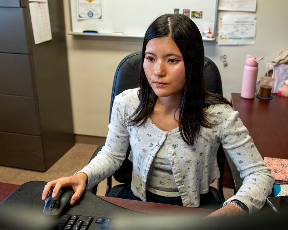 A young woman is seated at an office desk working at a computer, using a mouse and keyboard. She is wearing a light gray floral cardigan over a gray top and appears focused on a screen in front of her. The office setting includes a whiteboard, filing cabinet, bulletin board materials, and personal items on the desk.