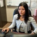 A young woman is seated at an office desk working at a computer, using a mouse and keyboard. She is wearing a light gray floral cardigan over a gray top and appears focused on a screen in front of her. The office setting includes a whiteboard, filing cabinet, bulletin board materials, and personal items on the desk.