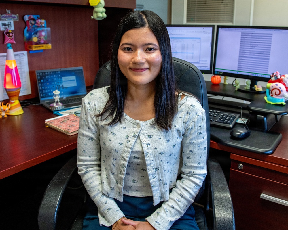 A young woman is seated in a black office chair at a wood-toned desk, smiling at the camera. The workspace features multiple monitors, a laptop, a lava lamp, and various decorative figurines and toys. The setting appears to be a personalized office or cubicle environment.