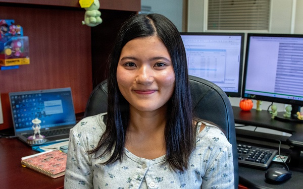 A young woman is seated in a black office chair at a wood-toned desk, smiling at the camera. The workspace features multiple monitors, a laptop, a lava lamp, and various decorative figurines and toys. The setting appears to be a personalized office or cubicle environment.