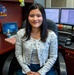 A young woman is seated in a black office chair at a wood-toned desk, smiling at the camera. The workspace features multiple monitors, a laptop, a lava lamp, and various decorative figurines and toys. The setting appears to be a personalized office or cubicle environment.