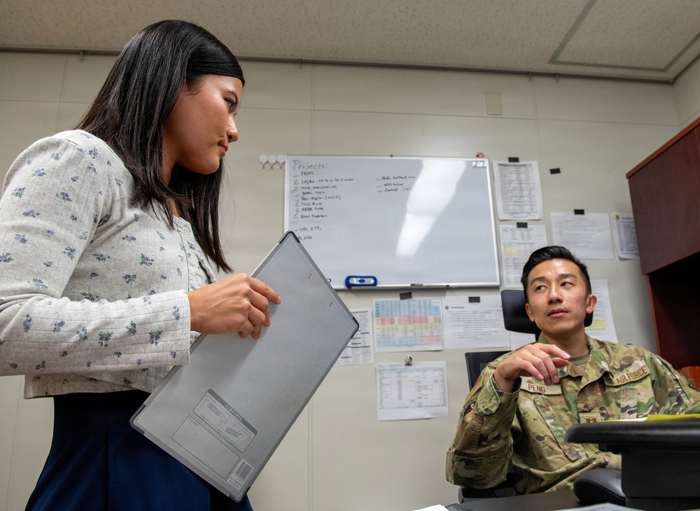 A civilian woman holding a tablet/clipboard is standing and speaking with a U.S. Air Force service member seated at a desk in an office environment. The military personnel member wears OCP (Operational Camouflage Pattern) uniform with a name tape reading 'PENG' and an Air Force patch. The setting appears to be a military administrative office with a whiteboard, posted documents, and office furniture visible in the background.