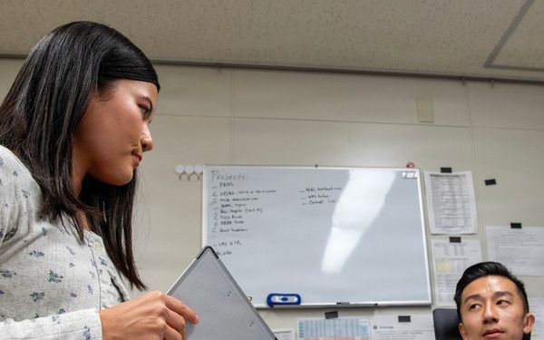 A civilian woman holding a tablet/clipboard is standing and speaking with a U.S. Air Force service member seated at a desk in an office environment. The military personnel member wears OCP (Operational Camouflage Pattern) uniform with a name tape reading 'PENG' and an Air Force patch. The setting appears to be a military administrative office with a whiteboard, posted documents, and office furniture visible in the background.
