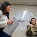 A civilian woman holding a tablet/clipboard is standing and speaking with a U.S. Air Force service member seated at a desk in an office environment. The military personnel member wears OCP (Operational Camouflage Pattern) uniform with a name tape reading 'PENG' and an Air Force patch. The setting appears to be a military administrative office with a whiteboard, posted documents, and office furniture visible in the background.