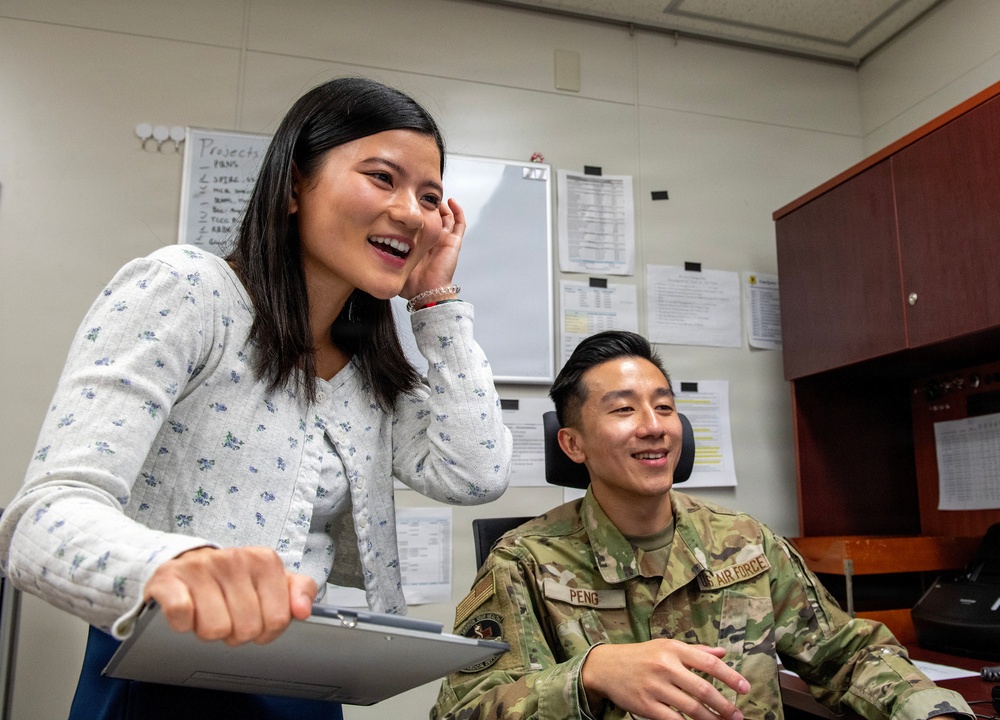 Two individuals are sharing a lighthearted moment in a military office setting. A civilian woman in a floral blouse is standing and laughing while holding a clipboard, alongside a seated U.S. Air Force servicemember in OCP camouflage uniform with the name tape 'PENG' visible. The setting appears to be a standard military administrative office with whiteboards, posted documents, and office furniture.