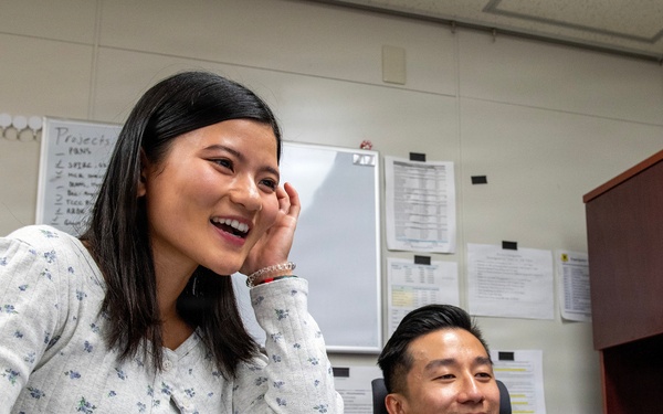 Two individuals are sharing a lighthearted moment in a military office setting. A civilian woman in a floral blouse is standing and laughing while holding a clipboard, alongside a seated U.S. Air Force servicemember in OCP camouflage uniform with the name tape 'PENG' visible. The setting appears to be a standard military administrative office with whiteboards, posted documents, and office furniture.
