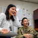 Two individuals are sharing a lighthearted moment in a military office setting. A civilian woman in a floral blouse is standing and laughing while holding a clipboard, alongside a seated U.S. Air Force servicemember in OCP camouflage uniform with the name tape 'PENG' visible. The setting appears to be a standard military administrative office with whiteboards, posted documents, and office furniture.