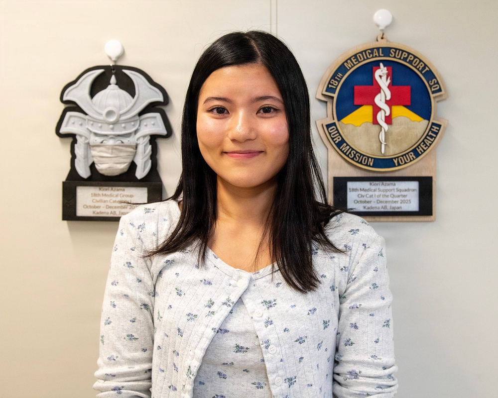 A young woman is posing for what appears to be an official recognition photo in front of two award plaques mounted on a wall. The plaques identify her as Kiori Azama, recognized by the 18th Medical Support Squadron at Kadena Air Base, Japan. She is smiling and dressed in a light floral cardigan.