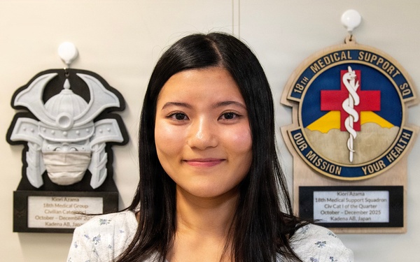 A young woman is posing for what appears to be an official recognition photo in front of two award plaques mounted on a wall. The plaques identify her as Kiori Azama, recognized by the 18th Medical Support Squadron at Kadena Air Base, Japan. She is smiling and dressed in a light floral cardigan.
