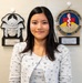 A young woman is posing for what appears to be an official recognition photo in front of two award plaques mounted on a wall. The plaques identify her as Kiori Azama, recognized by the 18th Medical Support Squadron at Kadena Air Base, Japan. She is smiling and dressed in a light floral cardigan.