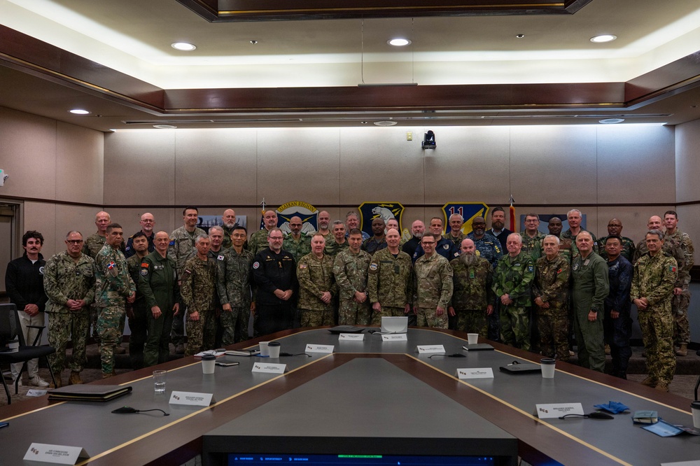 A large group of multinational military personnel pose for a formal group photograph in what appears to be a conference or briefing room. The individuals are wearing various military uniforms representing multiple nations, gathered around a large conference table. Military unit emblems/patches are visible on the wall behind them.