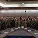 A large group of multinational military personnel pose for a formal group photograph in what appears to be a conference or briefing room. The individuals are wearing various military uniforms representing multiple nations, gathered around a large conference table. Military unit emblems/patches are visible on the wall behind them.