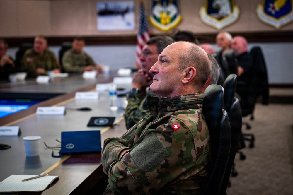 A military briefing or conference room setting showing multiple uniformed personnel seated around a large table. The foreground figure wears a camouflage uniform with a Swiss flag patch on the sleeve, suggesting an international military delegation. The room displays U.S. Air Force insignia and an American flag, indicating this is likely a U.S. Air Force installation hosting foreign military visitors.