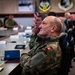 A military briefing or conference room setting showing multiple uniformed personnel seated around a large table. The foreground figure wears a camouflage uniform with a Swiss flag patch on the sleeve, suggesting an international military delegation. The room displays U.S. Air Force insignia and an American flag, indicating this is likely a U.S. Air Force installation hosting foreign military visitors.