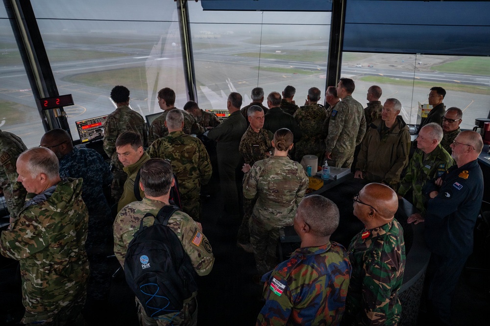 A large group of multinational military personnel are gathered inside what appears to be an air traffic control tower, visible by the panoramic windows overlooking an airfield. Personnel are wearing various national military uniforms and insignia, suggesting a multinational exercise or official visit. The group appears to be engaged in briefings or discussions around control tower equipment.