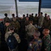 A large group of multinational military personnel are gathered inside what appears to be an air traffic control tower, visible by the panoramic windows overlooking an airfield. Personnel are wearing various national military uniforms and insignia, suggesting a multinational exercise or official visit. The group appears to be engaged in briefings or discussions around control tower equipment.