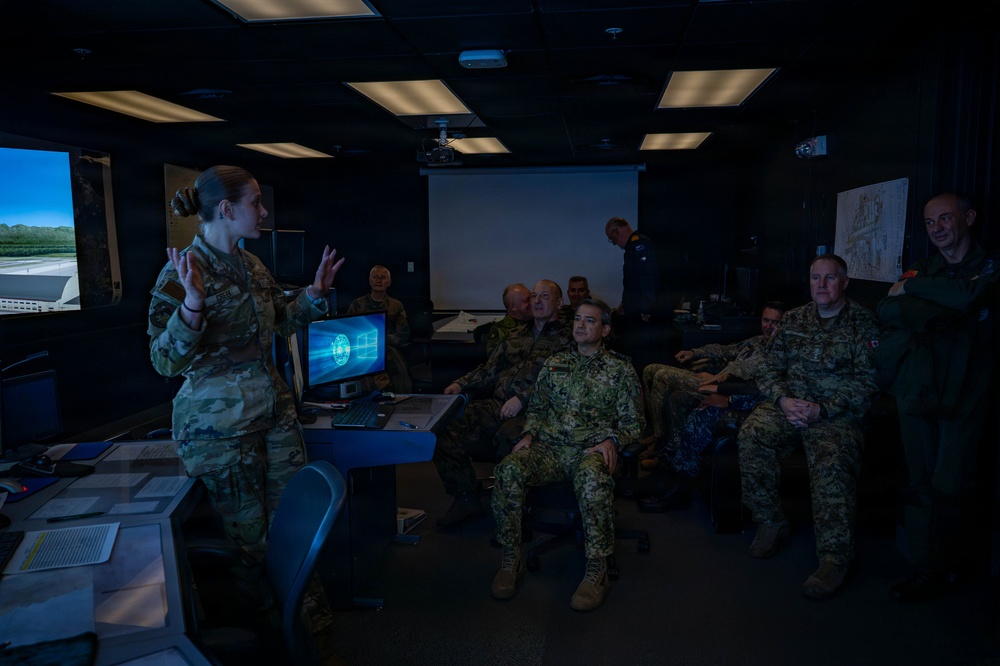 A female military service member in camouflage uniform is briefing a group of multinational military personnel in what appears to be a simulation or operations center. The room contains multiple screens, including a large display showing an aerial/runway view and a computer monitor with a digital interface. The audience consists of several officers in various military uniforms seated and attentively listening.