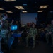 A female military service member in camouflage uniform is briefing a group of multinational military personnel in what appears to be a simulation or operations center. The room contains multiple screens, including a large display showing an aerial/runway view and a computer monitor with a digital interface. The audience consists of several officers in various military uniforms seated and attentively listening.