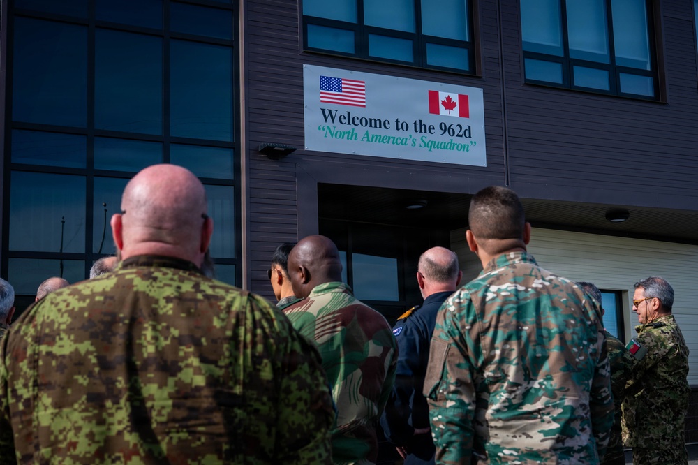 A group of military personnel in various uniforms are gathered outside a building displaying a welcome sign for the '962d North America's Squadron,' featuring both the American and Canadian flags. The individuals appear to be entering or assembling near the entrance of the facility. The scene suggests a multinational military visit or briefing event.