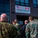 A group of military personnel in various uniforms are gathered outside a building displaying a welcome sign for the '962d North America's Squadron,' featuring both the American and Canadian flags. The individuals appear to be entering or assembling near the entrance of the facility. The scene suggests a multinational military visit or briefing event.