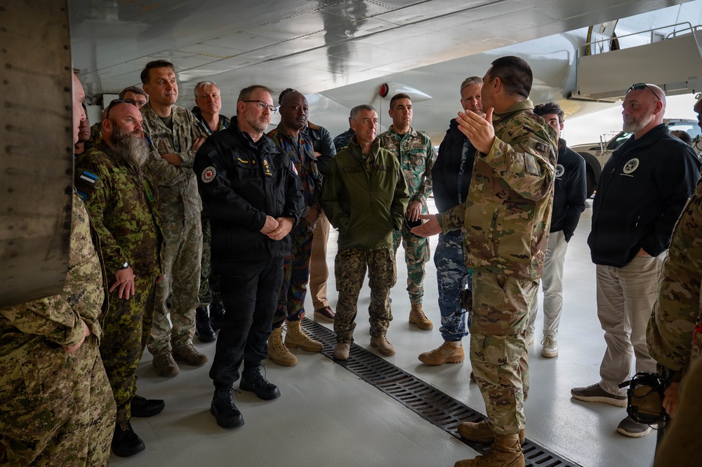 A military briefing is taking place inside what appears to be an aircraft hangar or under the wing of a large aircraft. A soldier in MultiCam/OCP camouflage uniform is addressing a diverse group of military and civilian personnel wearing various national uniforms and civilian attire. The gathering appears to be a multinational military delegation receiving an orientation or tour.
