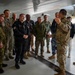 A military briefing is taking place inside what appears to be an aircraft hangar or under the wing of a large aircraft. A soldier in MultiCam/OCP camouflage uniform is addressing a diverse group of military and civilian personnel wearing various national uniforms and civilian attire. The gathering appears to be a multinational military delegation receiving an orientation or tour.