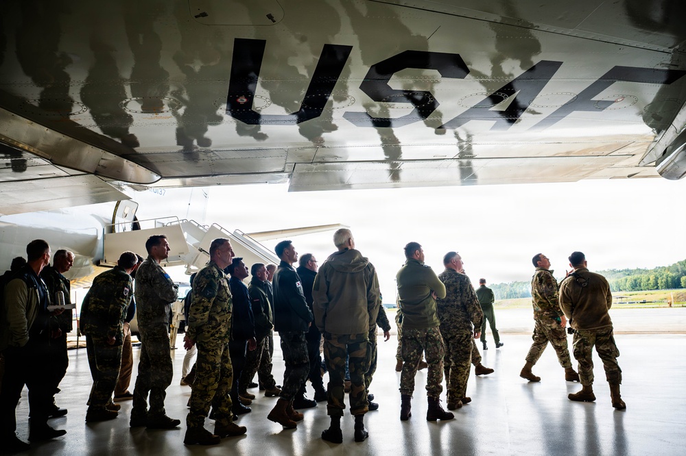 A group of military personnel in various camouflage uniforms are gathered beneath the fuselage of a large USAF aircraft inside or adjacent to a hangar. The underside of the aircraft prominently displays 'USAF' markings, and a boarding staircase is visible near the aircraft's entrance. The group appears to be receiving a briefing or tour of the aircraft.