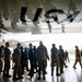 A group of military personnel in various camouflage uniforms are gathered beneath the fuselage of a large USAF aircraft inside or adjacent to a hangar. The underside of the aircraft prominently displays 'USAF' markings, and a boarding staircase is visible near the aircraft's entrance. The group appears to be receiving a briefing or tour of the aircraft.