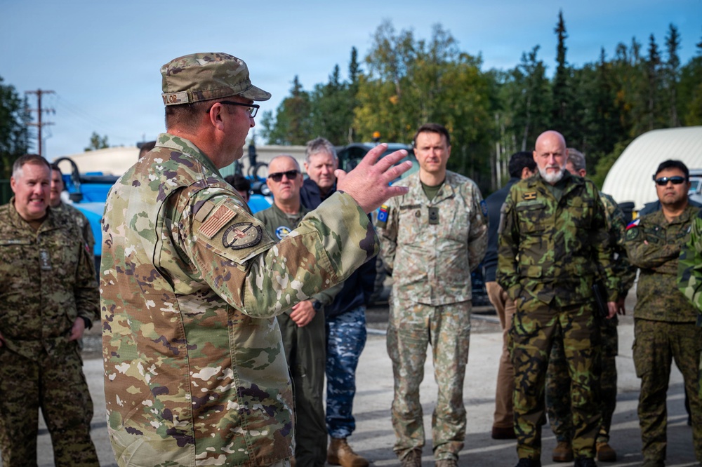 A U.S. military officer in OCP (Operational Camouflage Pattern) uniform with a U.S. flag patch is addressing a multinational group of military personnel outdoors. The gathering appears to be a briefing or orientation session, with personnel wearing various national military uniforms. The setting appears to be a military installation or staging area with vehicles and trees visible in the background.