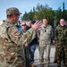 A U.S. military officer in OCP (Operational Camouflage Pattern) uniform with a U.S. flag patch is addressing a multinational group of military personnel outdoors. The gathering appears to be a briefing or orientation session, with personnel wearing various national military uniforms. The setting appears to be a military installation or staging area with vehicles and trees visible in the background.