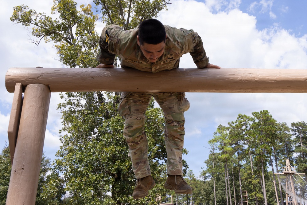 A U.S. Army soldier named Garcia is navigating an obstacle course, pulling themselves over a large horizontal log beam. The soldier is wearing OCP (Operational Camouflage Pattern) uniform with U.S. Army tape and rank insignia visible. The setting appears to be an outdoor military training facility surrounded by trees under a partly cloudy sky.