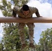 A U.S. Army soldier named Garcia is navigating an obstacle course, pulling themselves over a large horizontal log beam. The soldier is wearing OCP (Operational Camouflage Pattern) uniform with U.S. Army tape and rank insignia visible. The setting appears to be an outdoor military training facility surrounded by trees under a partly cloudy sky.