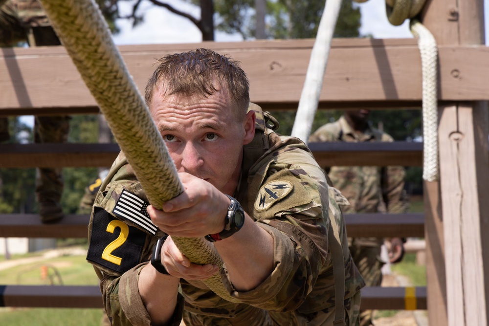 A U.S. Army soldier wearing MultiCam (OCP) camouflage uniform is climbing a thick rope on a military obstacle course. The soldier displays intense focus and physical exertion, gripping the rope tightly with both hands. A competitor number '2' armband and an American flag patch are visible on the left arm, suggesting this is a competitive military fitness event.