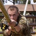 A U.S. Army soldier wearing MultiCam (OCP) camouflage uniform is climbing a thick rope on a military obstacle course. The soldier displays intense focus and physical exertion, gripping the rope tightly with both hands. A competitor number '2' armband and an American flag patch are visible on the left arm, suggesting this is a competitive military fitness event.