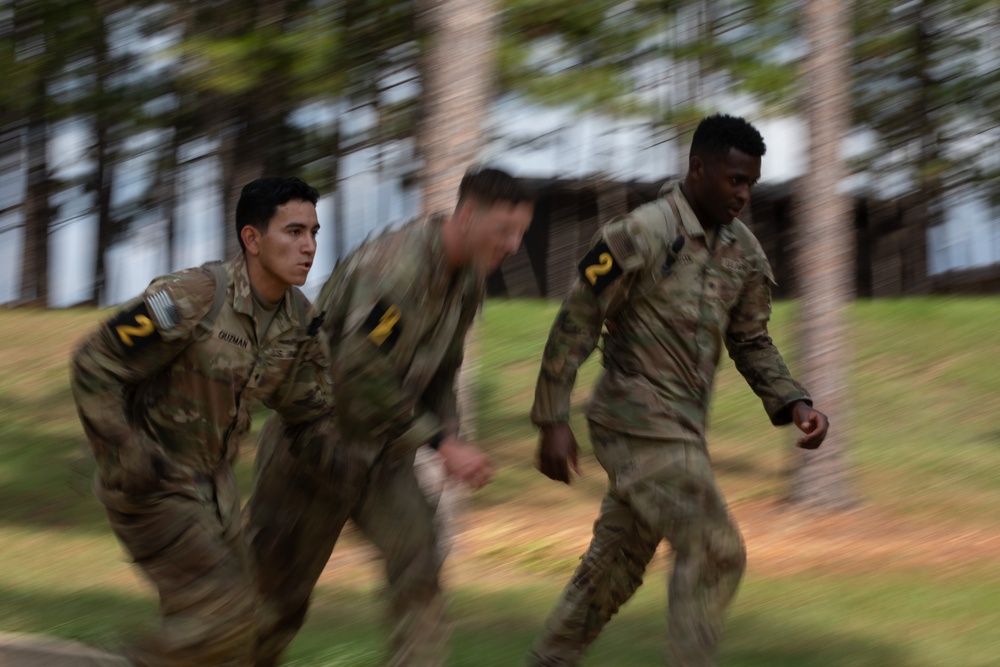 Three U.S. Army soldiers in MultiCam/OCP camouflage uniforms are running at speed in an outdoor wooded area. They wear shoulder patches with the number '2', indicating assignment to the 2nd Infantry Division or a similar unit. The image uses motion blur technique to convey speed and urgency.